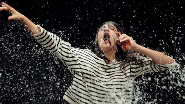 Young woman emotionally talking on mobile phone on black background. Gossips, talkative person. Water into face. Concept of emotions, communication, stress, unexpected news, ad