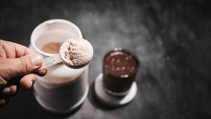Male hand holds measuring spoon with whey protein powder above plastic jar on dark background