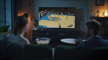 Back View of a Couple Spending Time at Home at Night, Sitting on a Sofa and Streaming a Live Basketball Playoffs Game on a TV in Their Apartment. Two Teams Playing the Match, Scoring Important Goals