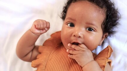 a close-up portrait of a small African-American baby girl in an orange bodysuit on the bed at home, a funny six-month-old black newborn baby lies on the back and smiles. View from above