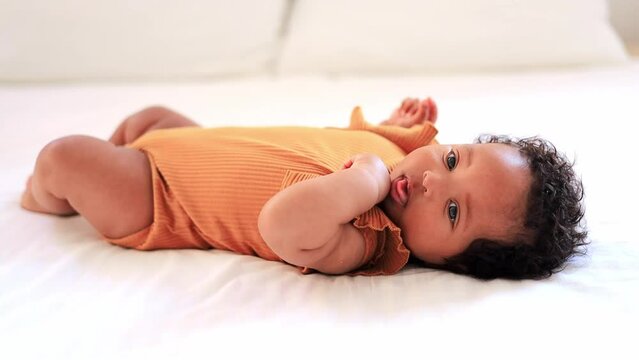 a close-up portrait of a small African-American baby girl in an orange bodysuit on the bed at home, a funny six-month-old black newborn baby lies on the back and smiles. View from above