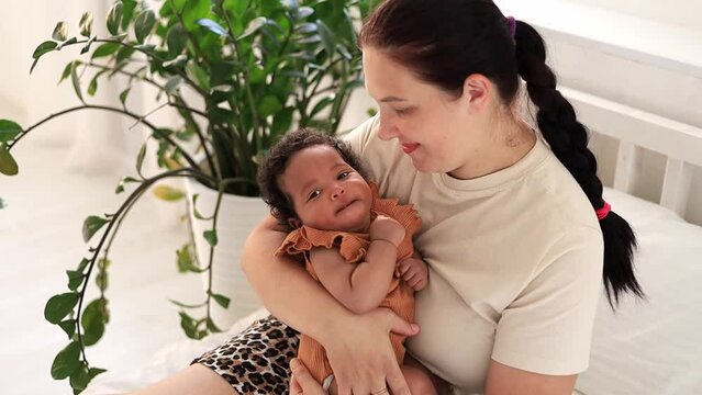 white Caucasian mom hugs and kisses an African-American black foster baby girl at home on the bed, maternal love and care, a mother of European appearance with a black child in her arms