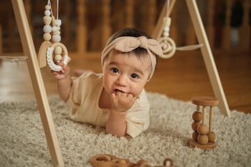Exploring the World of Baby Teething: A one-year-old cherub lays on a beige mat, gnawing her fingers to relieve teething discomfort, surrounded by wooden toys