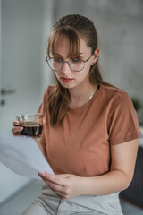 Young female student drinking coffee  and looking at paper, learning at home concept