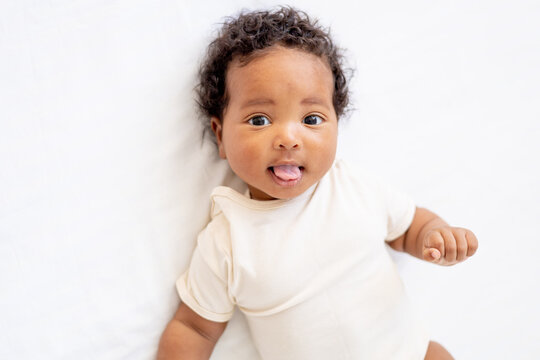 close-up portrait of a small African American baby girl in a white bodysuit on a cotton bed at home, funny six-month-old smiling joyful black newborn baby shows tongue