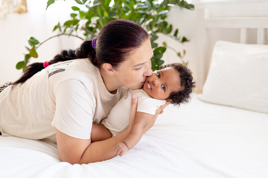 white Caucasian mom hugs and kisses an African-American black foster baby girl at home on the bed, maternal love and care, a mother of European appearance with a black child in her arms