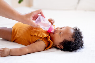 Obraz na płótnie Canvas a small African-American baby girl drinks water or milk from a bottle in an orange bodysuit on a white bed, her mother's hand feeds a six-month-old black newborn baby from a bottle