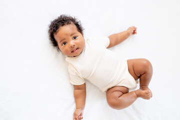 little african American baby girl in a white bodysuit on a cotton bed at home, six-month-old...