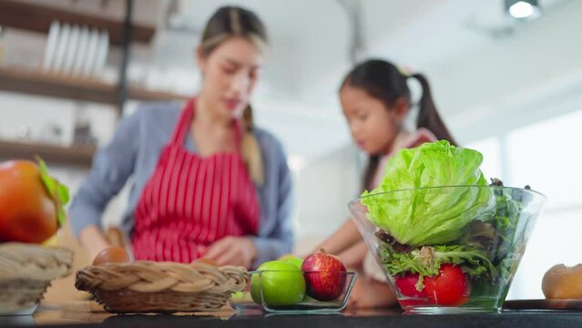 Close up fresh vegetables and fruits in glass bowl on table in kitchen room and background mother and daughter enjoy preparation of vegan salad. Healthy food concept