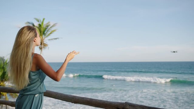 Drone shoot blonde woman video blogger talking on camera on tropical island with ocean view. Female vlogger making video on drone in Sri Lanka from back view. Traveler filming movie in slow motion.