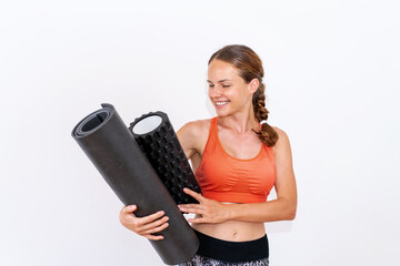 studio shot female athlete holding exercise equipment foam roller rolled yoga mat and smiling, wearing sportswear, in front of white background.