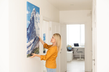 Canvas print with gallery wrap interior decor. Woman hangs landscape photography on white wall. Hands holding photo canvas print