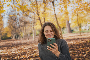 Portrait of young woman in a sweater with a mobile phone in her hands sitting in an autumn park