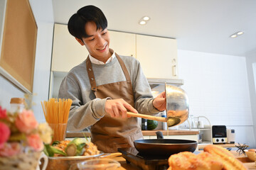Happy young Asian man pouring pancake batter into a hot frying pan, preparing breakfast in cozy kitchen