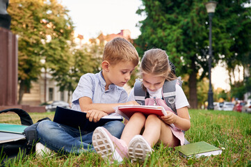 Fototapeta premium Sitting on the green grass. Young school children of boy and girl are together outdoors