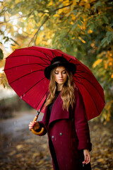 Close up portrait of a Beautiful girl in dark dress 
and black hat standing near colorful autumn leaves. Art work of romantic woman .