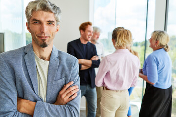 Smiling businessman standing with arms crossed wearing blazer against team discussing at workplace