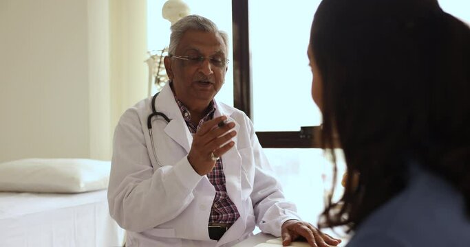 Senior Indian male GP doctor in white uniform consult female patient in private clinic. Experienced older therapist talks to woman client during professional medical consultation in hospital. Medicine
