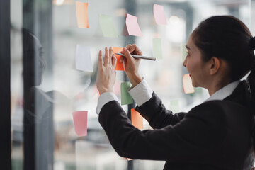 A millennial Asian businesswoman shares ideas using sticky notes, which she places on a glass wall.