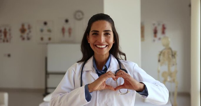 Portrait smiling positive Latina female cardiologist in white medical uniform pose at workplace showing heart shape with joined folded fingers. Cardiology, cardiovascular disease prevention, medicine