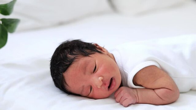 portrait of a newborn black African-American baby with open eyes, a small dark-skinned baby close-up lying on the bed on his tummy in the bedroom waking up or falling asleep in a white bodysuit