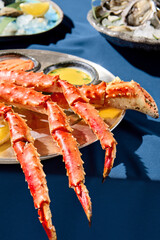Closeup of a gourmet seafood dish featuring prepared crab phalanges with lemon, sauces, and fresh oysters on ice. Presented on a metal dish on a blue tablecloth with a stark blue background