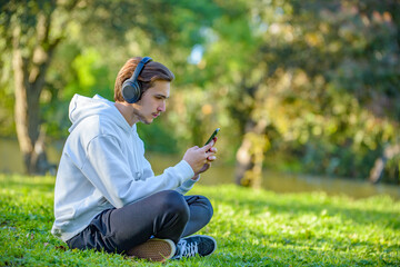 Naklejka na ściany i meble Portrait of young man in headsphone with a phone in his hands outdoors. Young guy looks at the screen of a cell phone while sitting on green lawn in the park. White man with smartphone outdoors