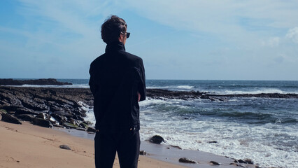 Young man stands on the empty sandy beach on the summer day and contemplating ocean water surface....