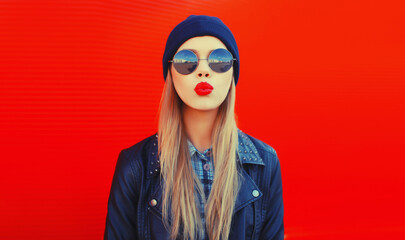Portrait of stylish beautiful blonde woman blowing her red lips sending sweet air kiss wearing round sunglasses, black rock style, jacket and hat - Powered by Adobe
