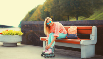 Stylish blonde young woman on roller skates with backpack posing in city park