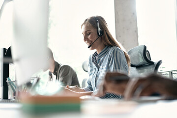 Friendly smiling woman call center operator with headset using computer at office.