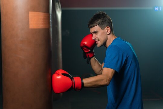 Tall white man in a blue tee shirt engages in a focused boxing punching exercise with a heavy bag, showcasing his dedication to boxing and fitness in a boxing gym