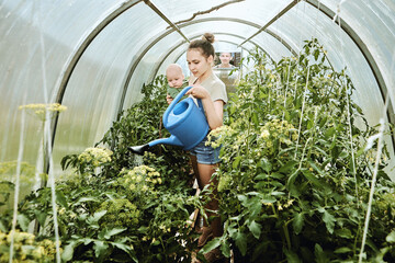 Young woman holds baby in one hand and holds watering can in other while tending plants in...