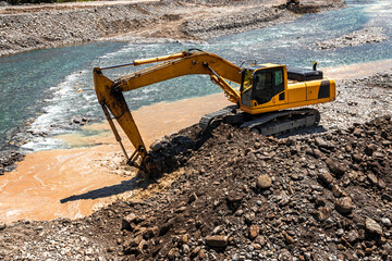 The excavator is working on expanding the riverbed. A yellow excavator at a construction site near...