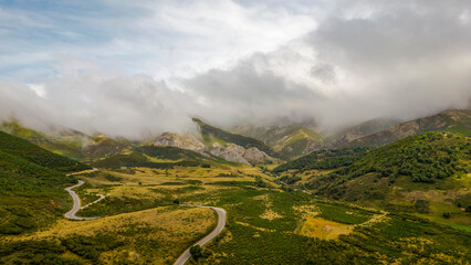 una carretera serpentea entre montañas nubladas, rodeada de verdes prados con las altas montañas del pirineo al fondo y un cielo azul nublado - obrazy, fototapety, plakaty