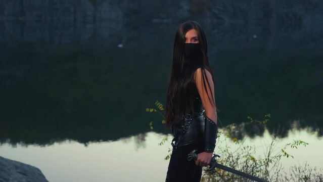 Danger fantasy woman warrior assassin holds dagger in hands walks at night nature. Rear view, looking back armed girl. Black leather costume, hood. Ninja soldier with sword knife. river rocks night 4k