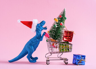 Happy blue dinosaur in Santa Claus hat carrying shopping cart with Christmas tree and gift boxes on pastel pink background.