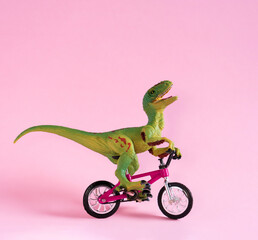 Fototapeta premium Cute happy dinosaur toy riding bicycle on pastel pink background. Cute eco friendly transport concept card.