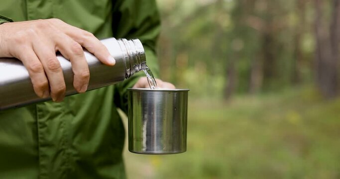 man pours hot tea from thermos flask into a mug in forest. nature adventure, tourism and camping concept
