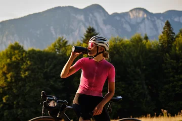 Foto op Canvas Cyclist silhouette. Female cyclist in cycling clothes and a helmet drinks water from a sports bottle at sunset. © Ketrin