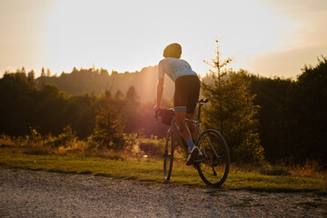 A fit male cyclist practicing on a gravel road at sunset. He is riding a gravel bike with a view of...