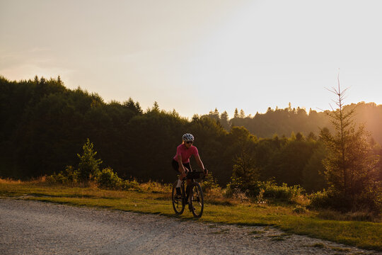 A fit female cyclist, wearing a cycling kit and helmet, is riding a gravel bike on a gravel road at sunset. Sports motivation image.