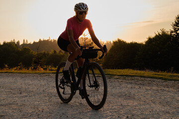A female cyclist, wearing a pink cycling jersey and a white helmet, is riding a bicycle on a gravel road at sunset.Woman cycling in the nature. Active lifestyle concept. Cycling gravel adventure.