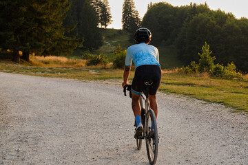 Naklejka premium A fit male cyclist practicing on a gravel road at sunset. He is riding a gravel bike with a view of the mountains. Bucegi Mountains, Romania