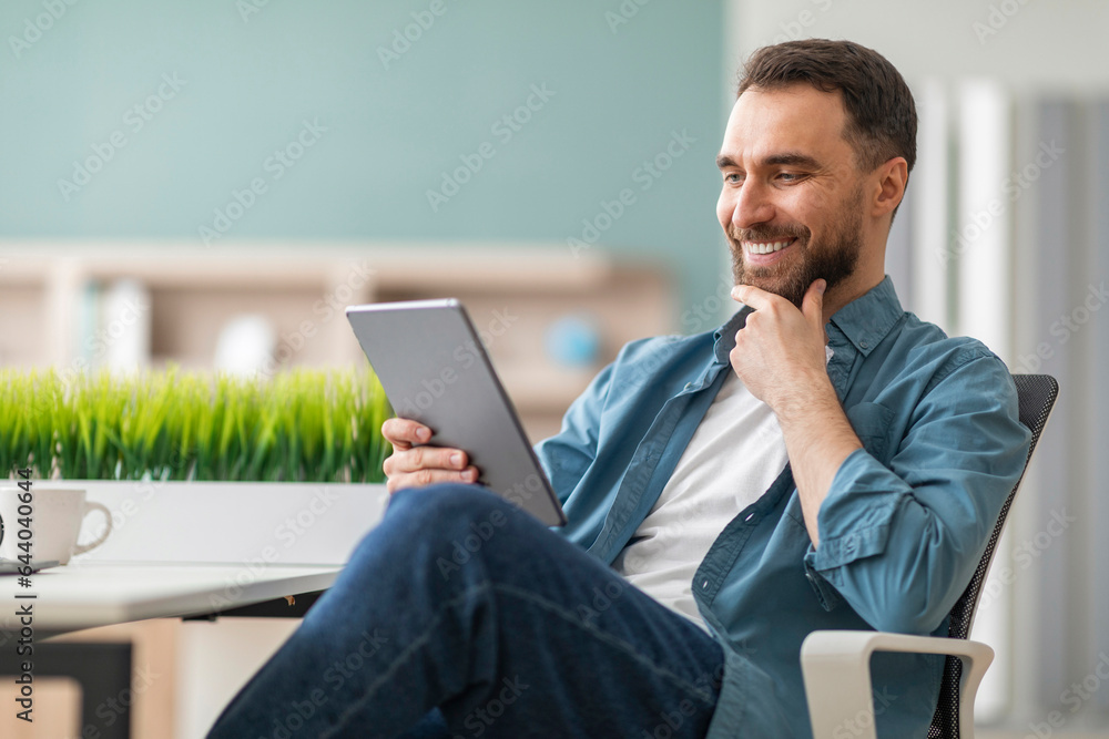 Poster smiling young man using digital tablet while sitting at desk at office