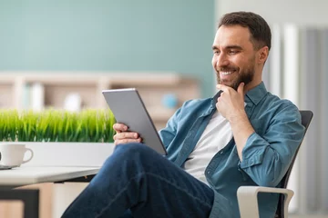 Gordijnen Smiling Young Man Using Digital Tablet While Sitting At Desk At Office © Prostock-studio