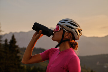 Cyclist silhouette. Female cyclist in cycling clothes and a helmet drinks water from a sports bottle at sunset. - obrazy, fototapety, plakaty