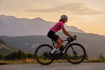 Woman cyclist is riding a gravel bike on a gravel road  at sunset with a view of the...