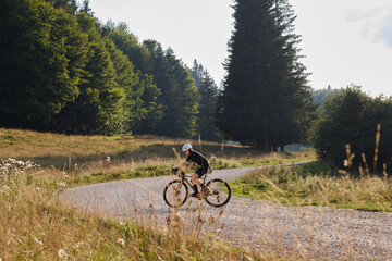 Woman cyclist wearing black cycling kit. She is riding a gravel bike on a gravel road at sunset with a view of the mountains.Empty mountain road.Cycling gravel adventure in Romania.Bucegi Natural Park