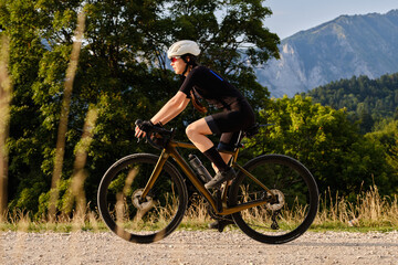 Obraz premium Woman cyclist wearing black cycling kit. She is riding a gravel bike on a gravel road at sunset with a view of the mountains.Empty mountain road.Cycling gravel adventure in Romania.Bucegi Natural Park